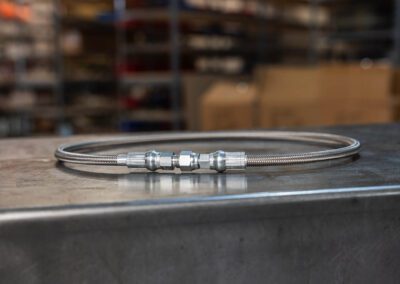 Braided stainless steel Teflon hose with metal fittings, shown on a metal workbench at PT Hose & Bearing.